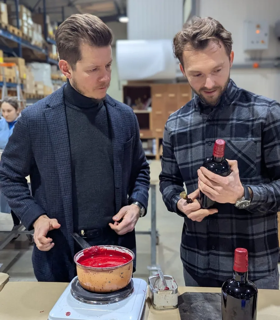 Deux hommes dans un atelier d'embouteillage examinent une bouteille à côté d'un pot de cire rouge sur une balance.