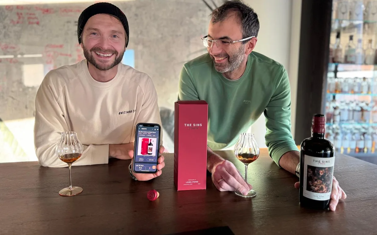 Die beiden Distilia-Gründer an einem Verkostungstisch, auf dem Handy die RumX-App, daneben Flasche und Box von The Sins Avarice.