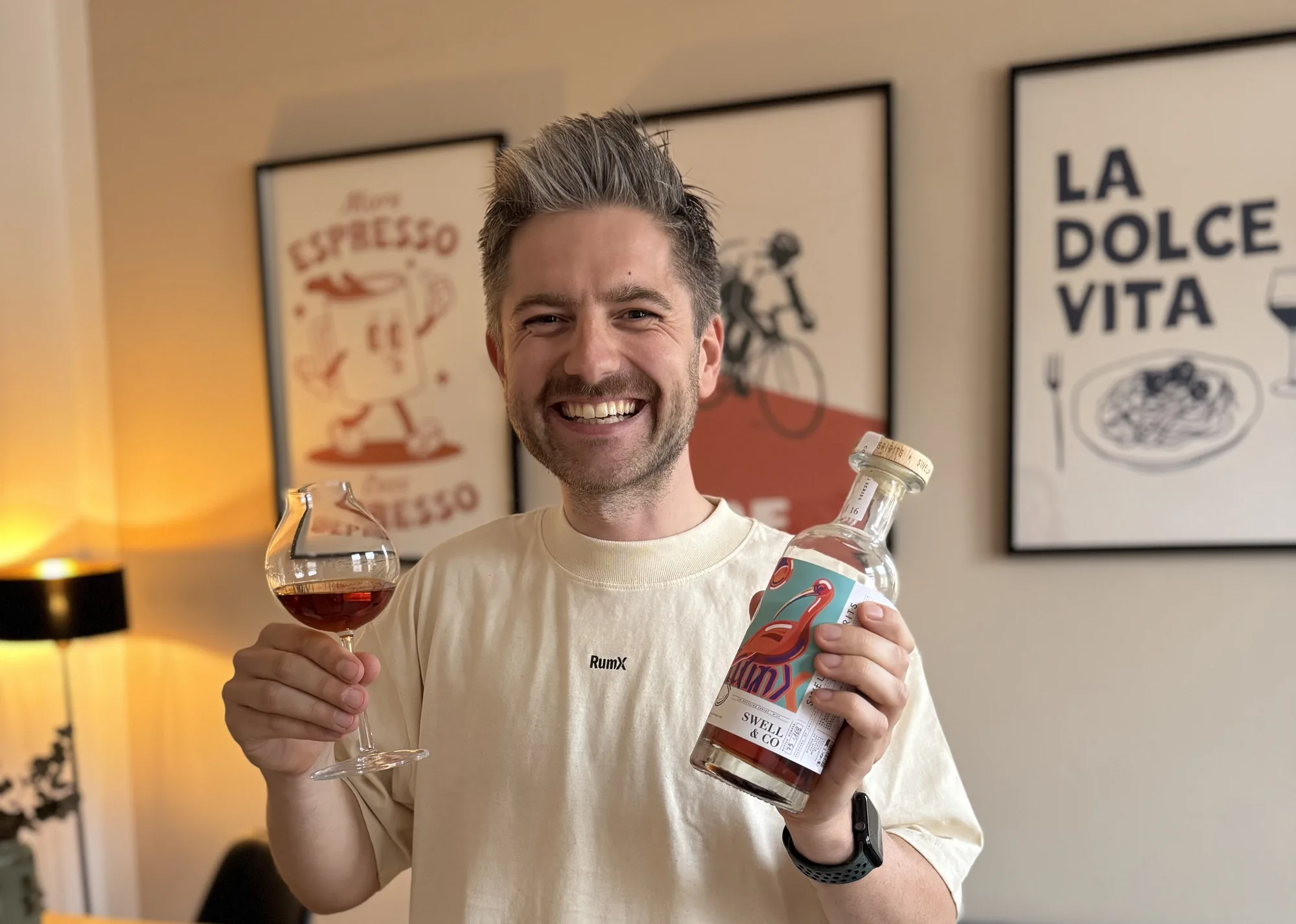 Oliver Gerhardt en t-shirt RumX, souriant face à la caméra, un verre de rhum et une bouteille en main.