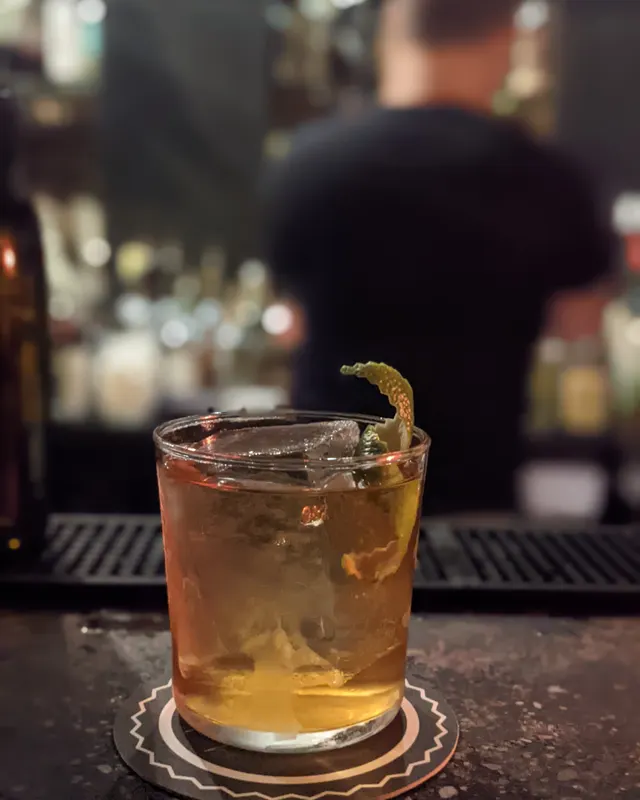 Old Fashioned mit Orangenzeste auf dunklem Bartresen, Barkeeper unscharf im Hintergrund