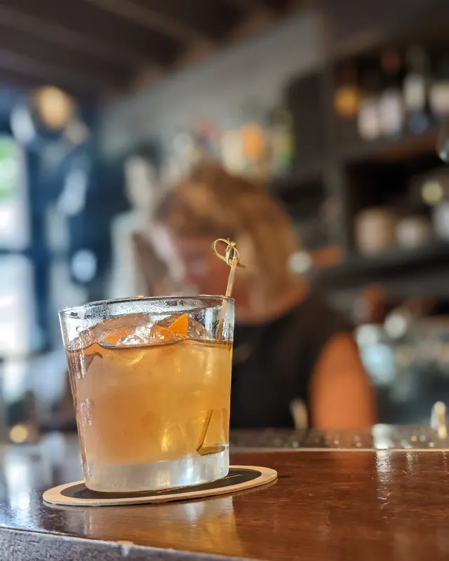 Old Fashioned mit Orangengarnitur am Spieß vor beleuchtetem Barregal