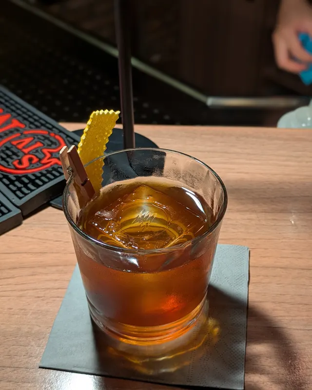 Dunkler Old Fashioned mit gelber Garnierklammer auf dunklem Holztresen